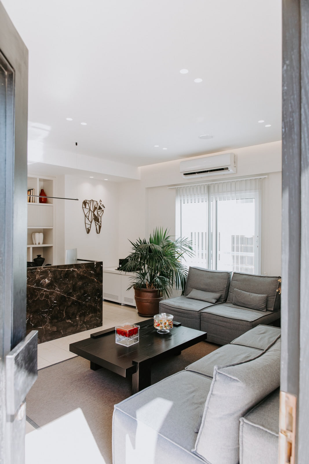 The waiting room through the doorway, with grey sofas and a kentia palm