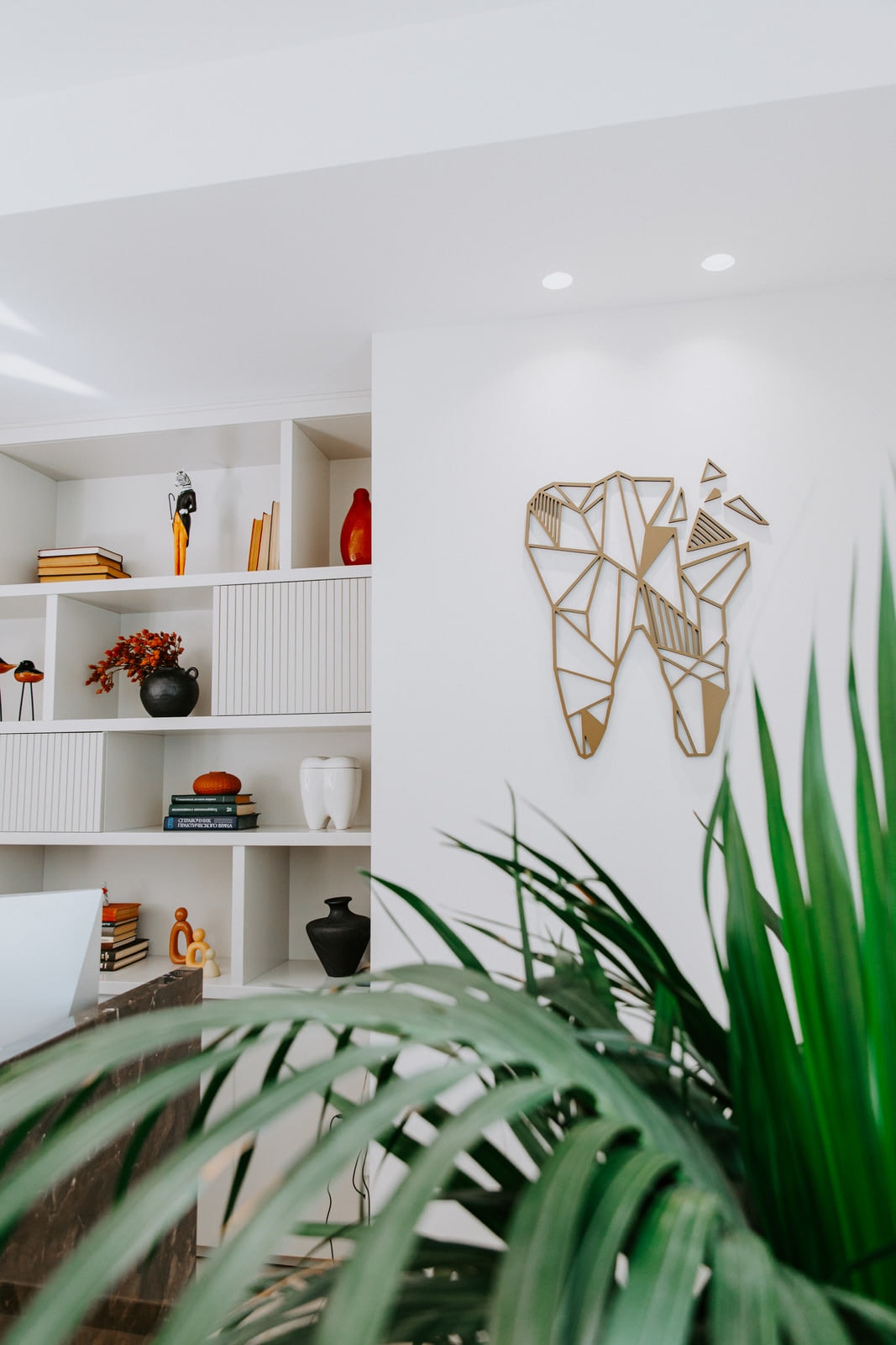The brass geometric tooth above the white shelving, seen past a palm